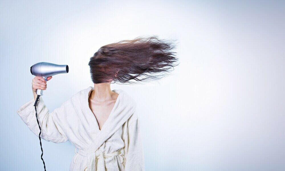 A white baqckground with a light skinned lady blow drying her brunette hair. Her hair is covering her face in accordance with the blow dryer.