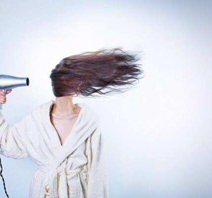 A white baqckground with a light skinned lady blow drying her brunette hair. Her hair is covering her face in accordance with the blow dryer.