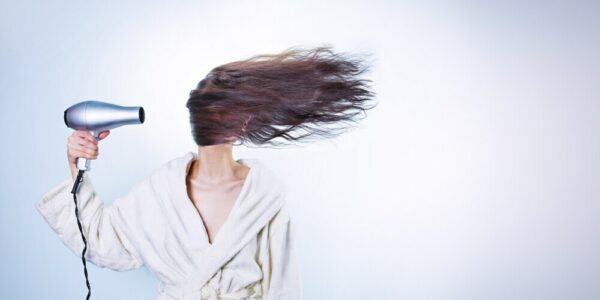 A white baqckground with a light skinned lady blow drying her brunette hair. Her hair is covering her face in accordance with the blow dryer.